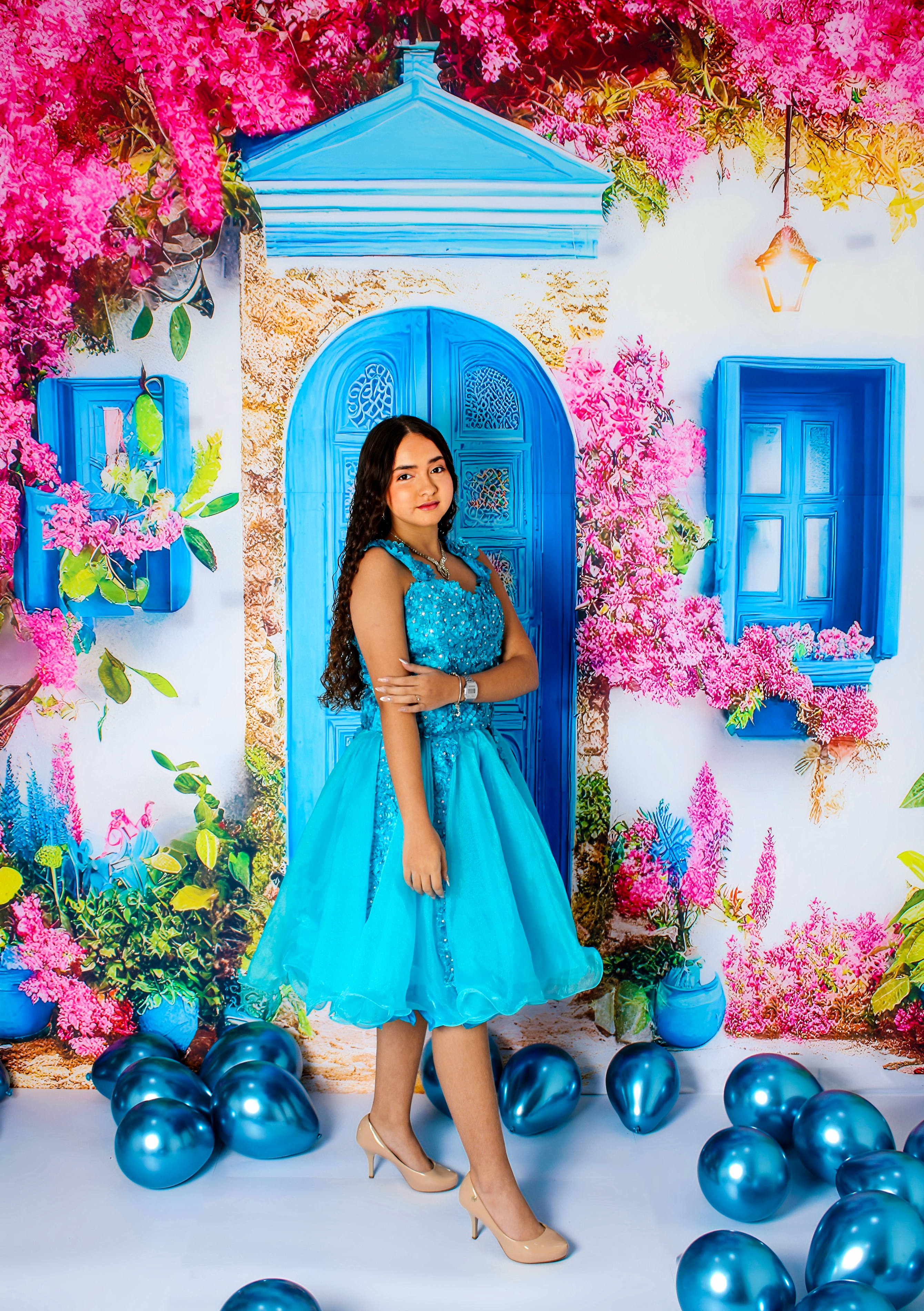 Jovem com vestido curto azul-turquesa em pé, rodeada de balões azuis, comemorando seus 15 anos.