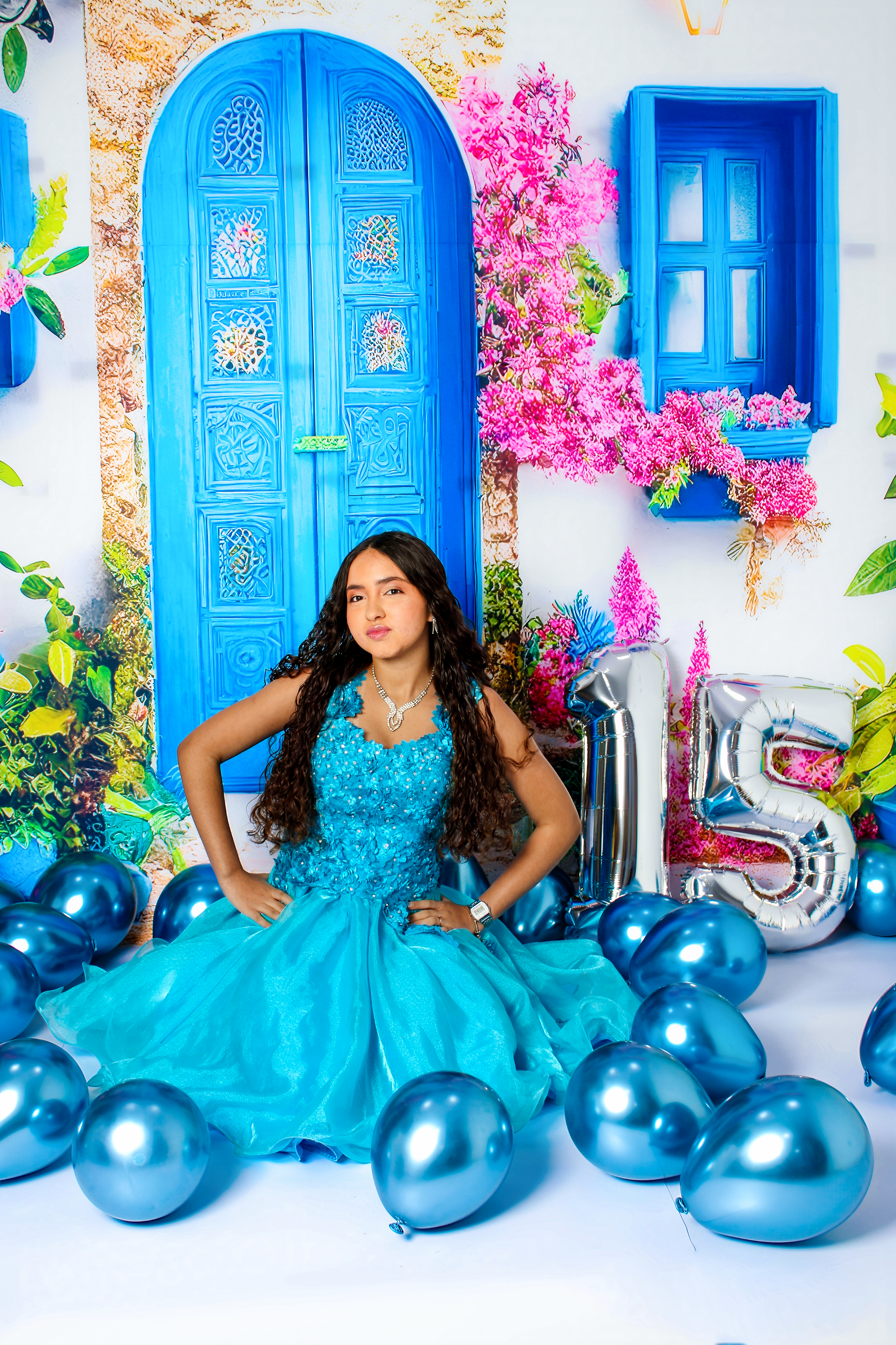 Jovem usando vestido longo azul-turquesa, rodeada de balões e flores cor-de-rosa em cenário de 15 anos.