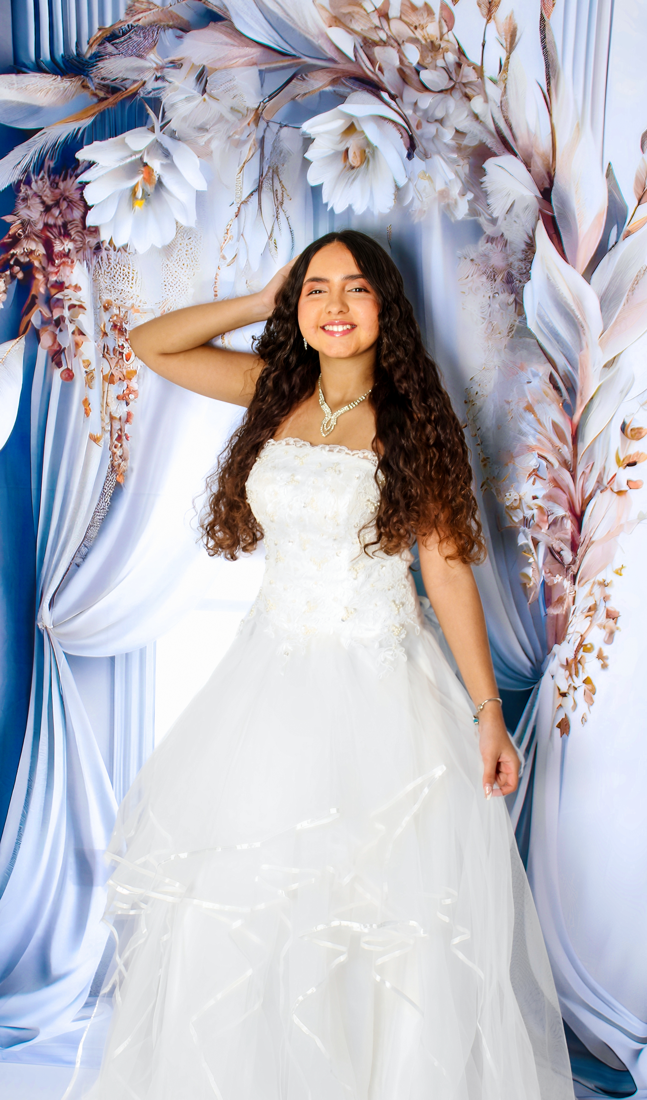 Jovem usando vestido de festa branco e longo, posando em frente a uma porta azul e flores cor-de-rosa para seus 15 anos.