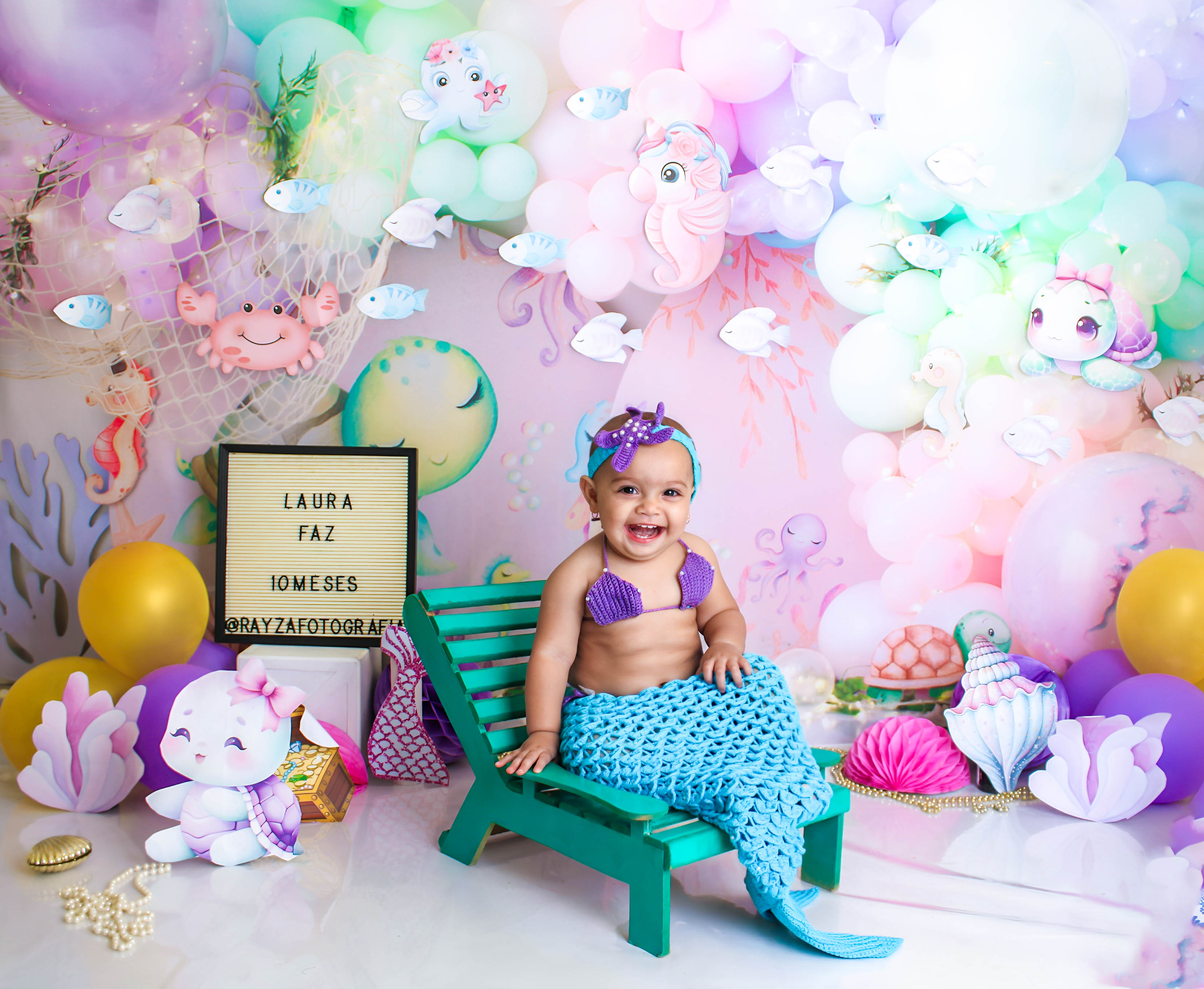 Bebê fantasiada de sereia, com rabo e top colorido, posando em um cenário decorado com balões e conchas.