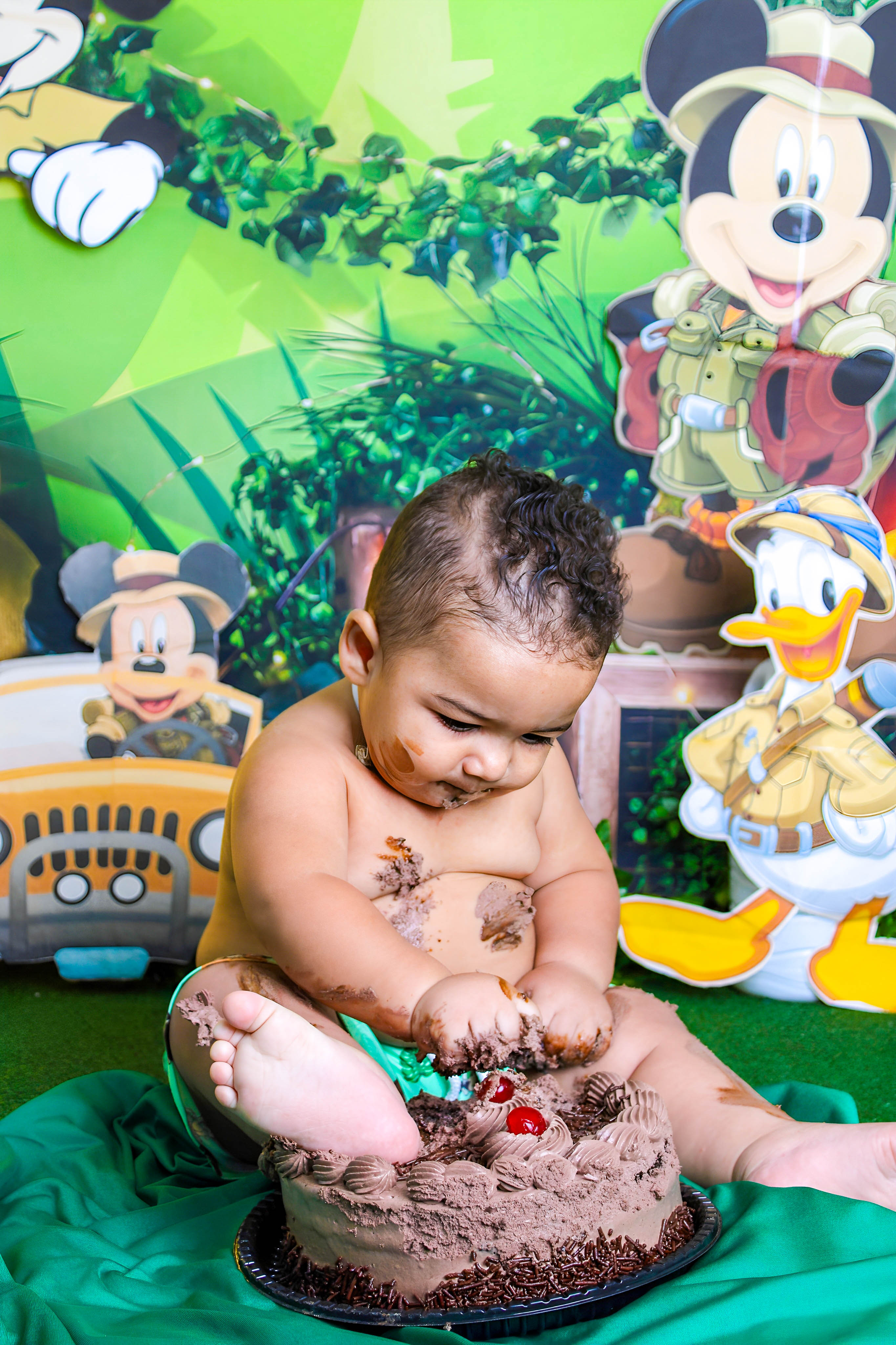 Bebê sem camisa, interagindo com bolo de chocolate em um ensaio Smash the Cake inspirado no Mickey Mouse.