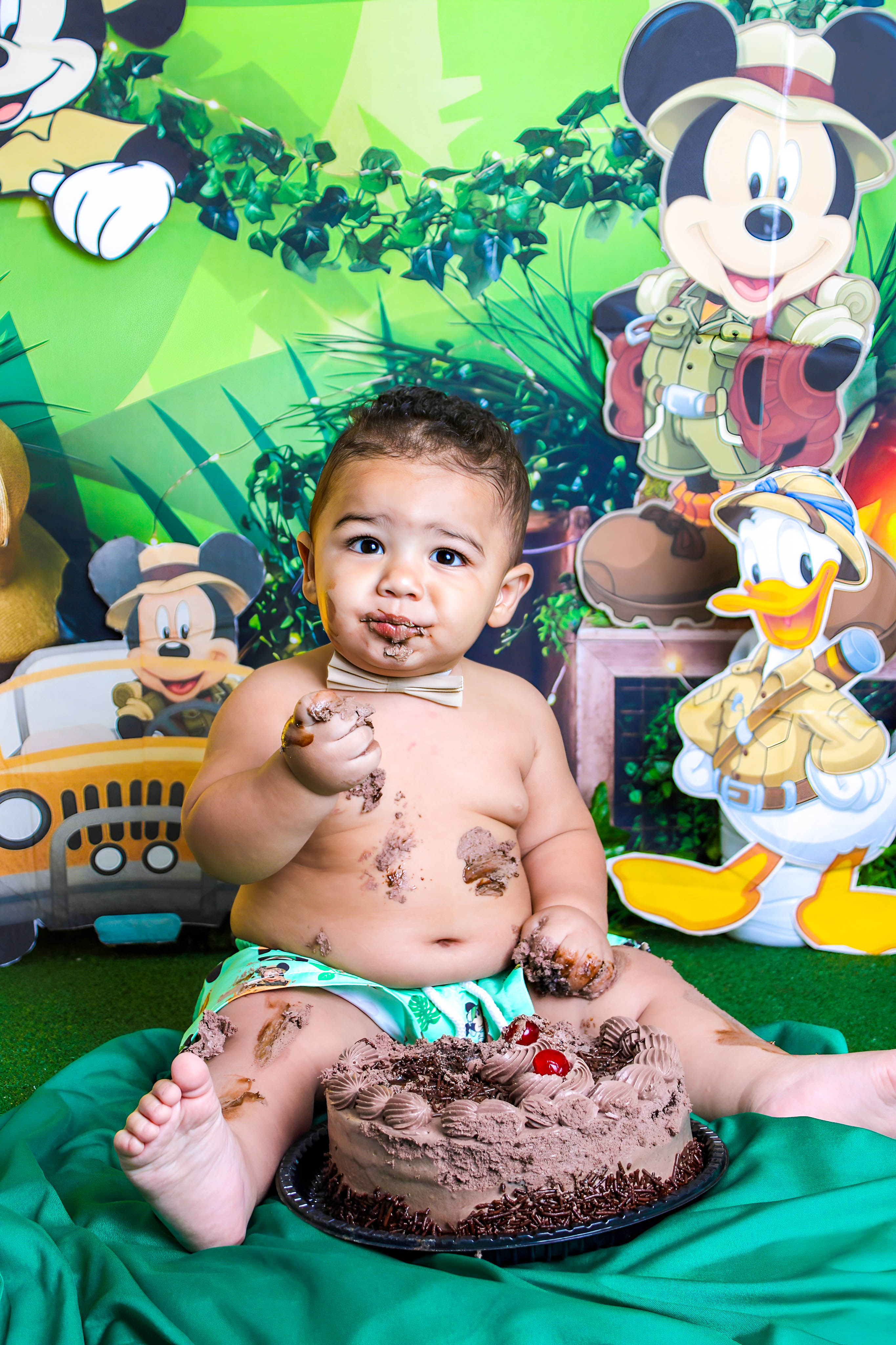 Bebê lambuzado de chocolate, sorrindo durante o ensaio Smash the Cake com cenário do Mickey.