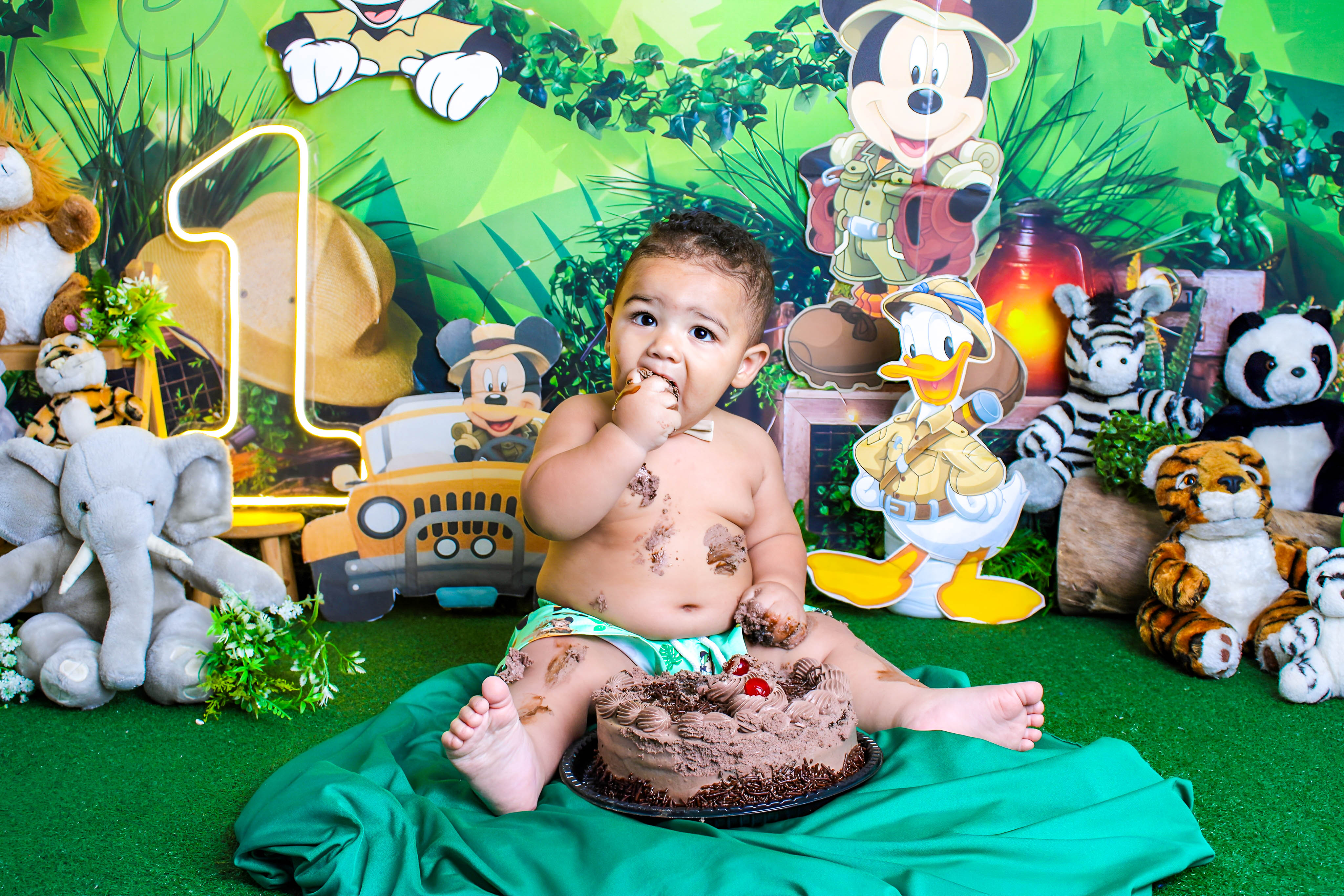 Bebê explorando o bolo de chocolate com as mãos em um cenário repleto de itens do Mickey Mouse.