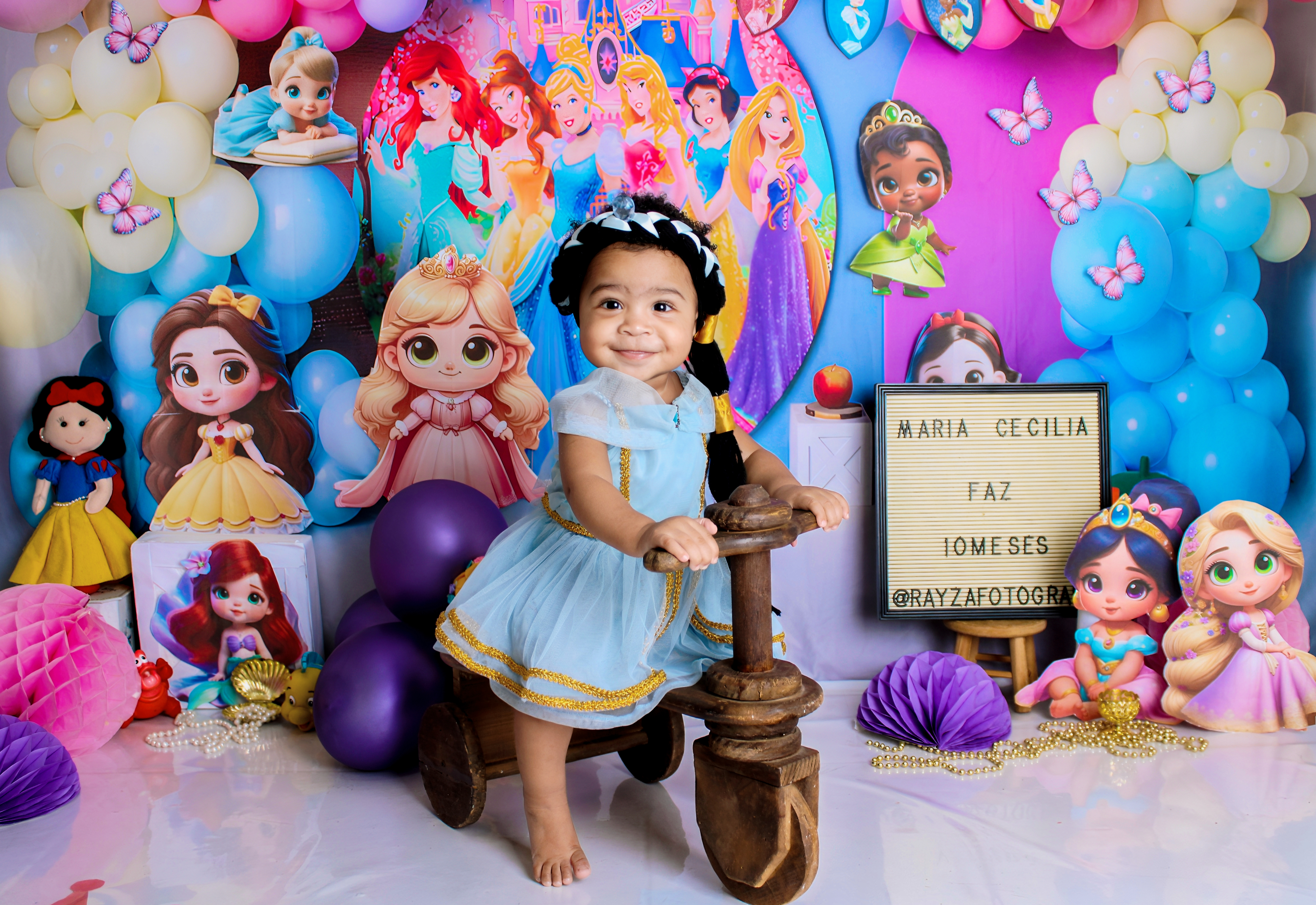 Bebê com vestido lilás, sentada em uma poltrona, em um ensaio temático rodeado de balões e figuras lúdicas.