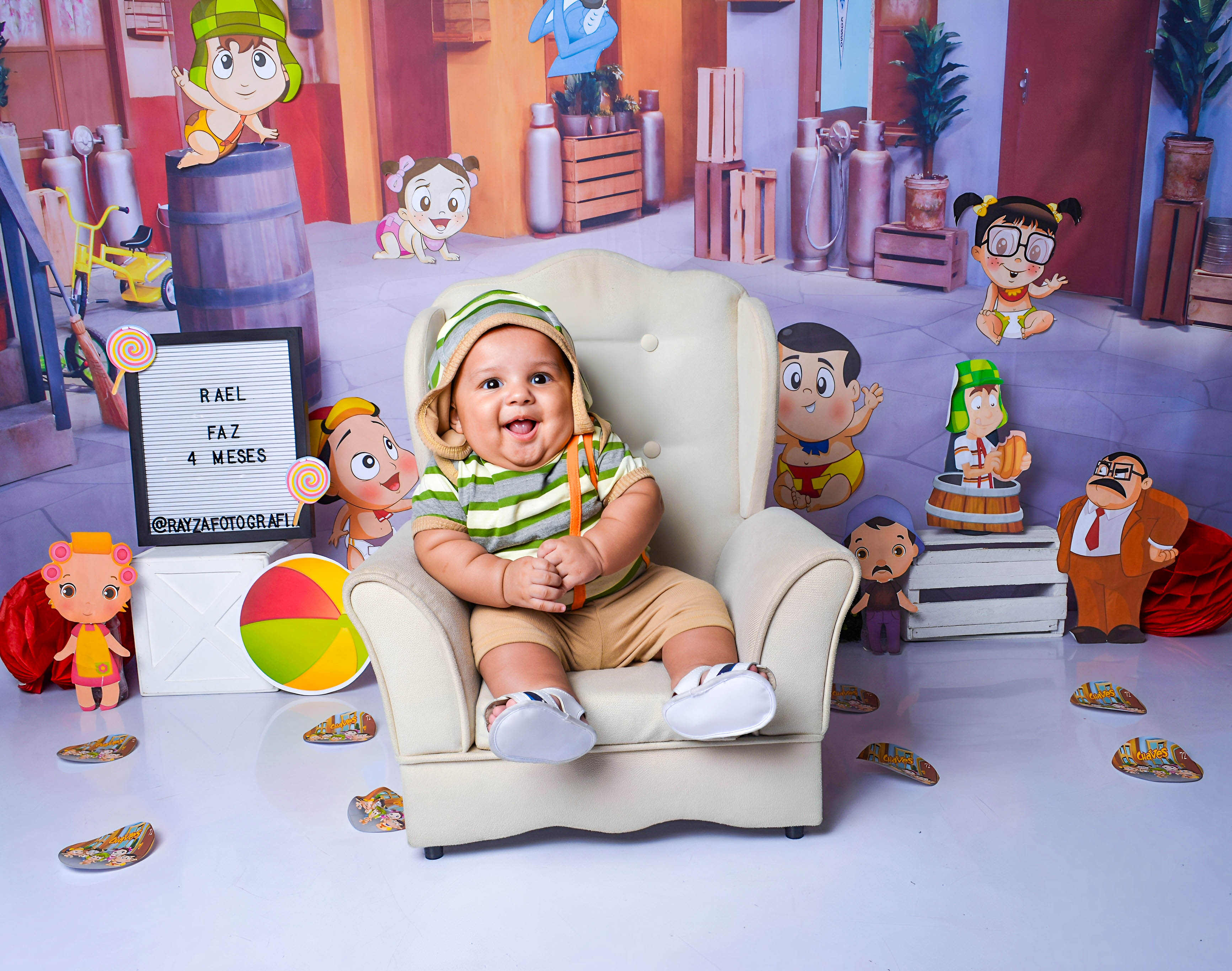 Bebê sentado em uma poltrona cinza, vestindo touca e figurino listrado, em um cenário infantil repleto de balões.