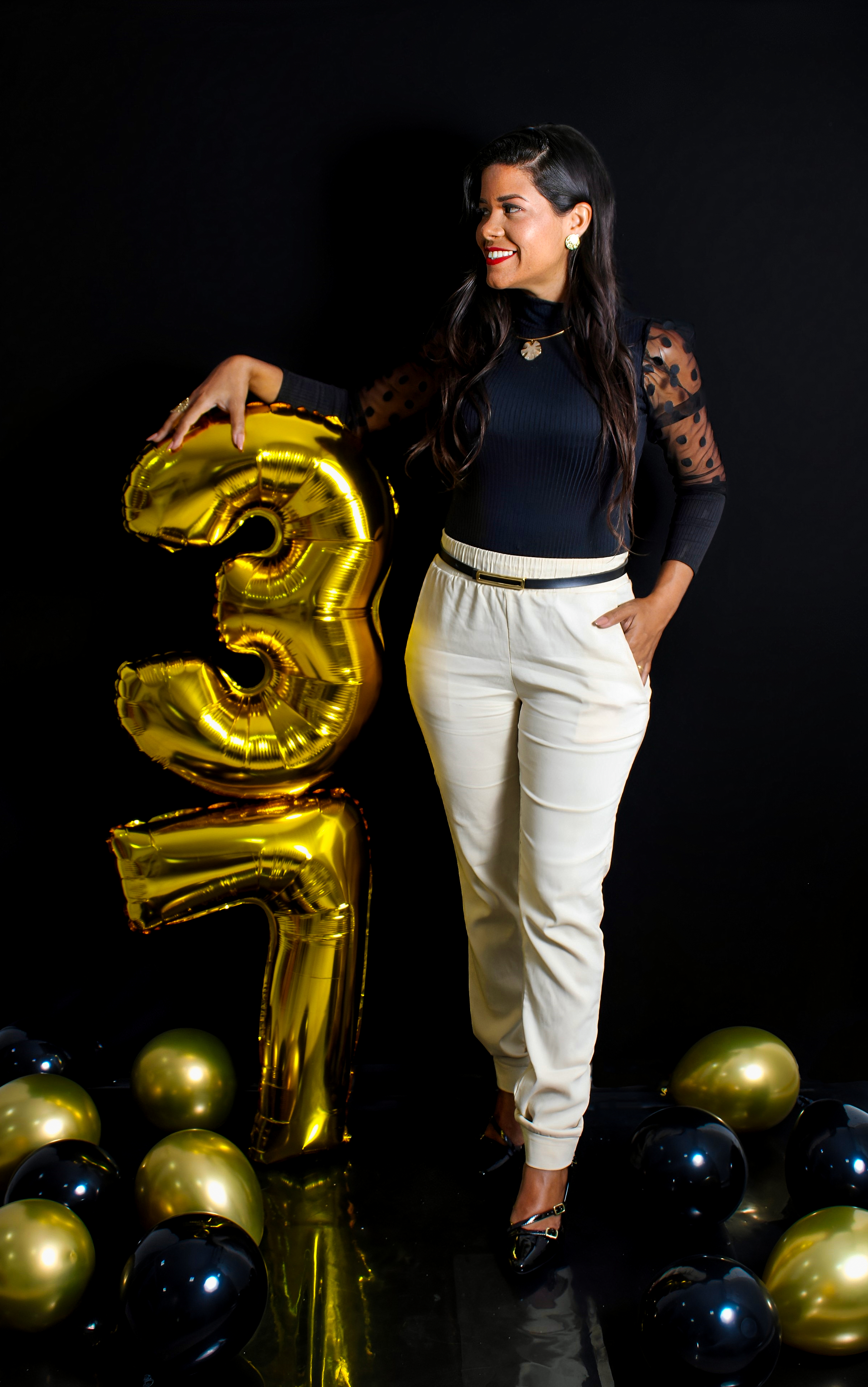 Mulher em pé ao lado de um balão dourado em formato de 3, usando roupa preta e calça clara, em cenário com fundo escuro.