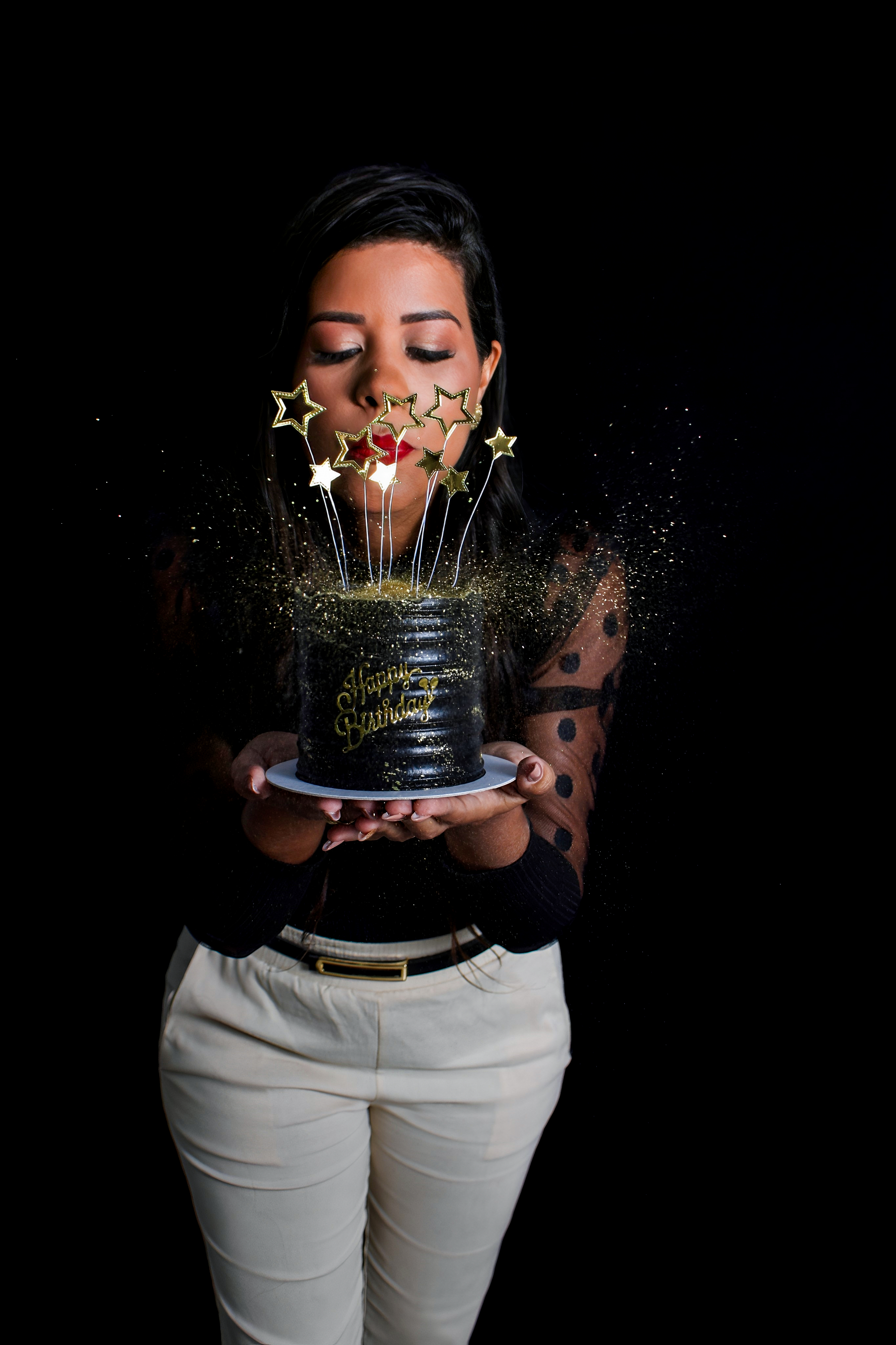 Mulher segurando um bolo decorado com coroa dourada, posando em um cenário de aniversário com balões pretos e dourados.