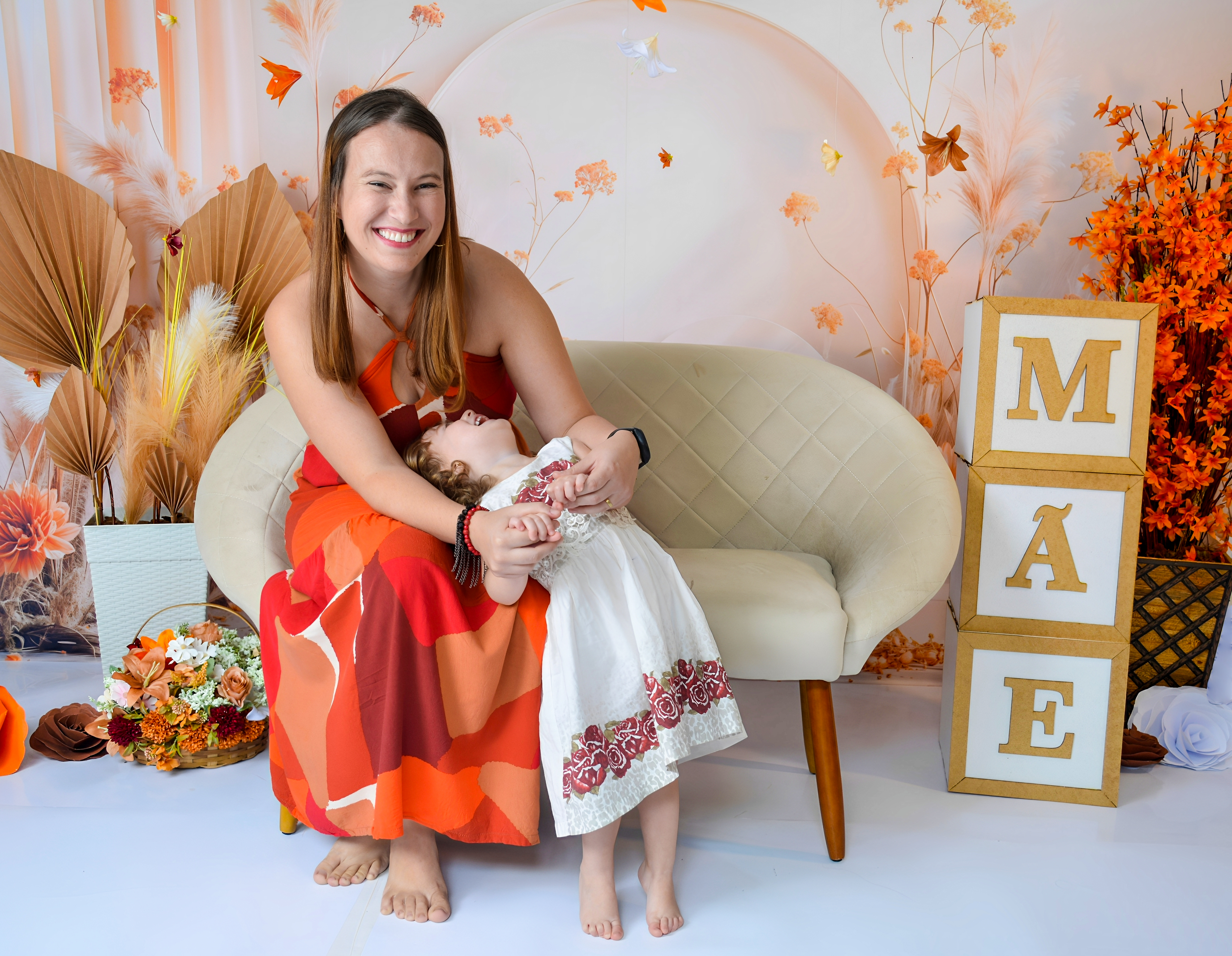 Mãe segurando a filha no colo, ambas com vestidos laranja e branco em um ensaio fotográfico.