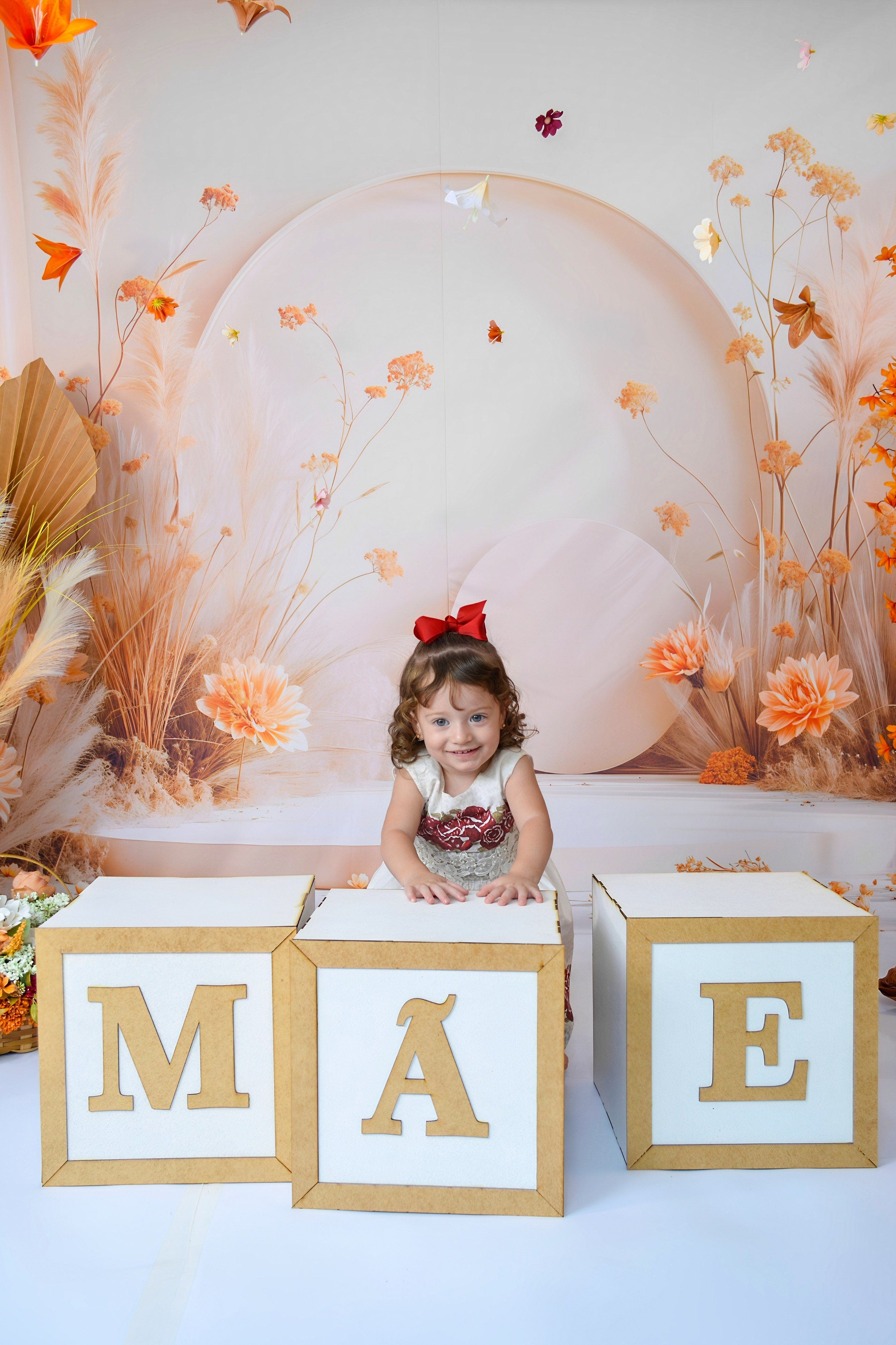 Menina com vestido laranja e branco posando diante de caixas que formam a palavra 'MÃE'.