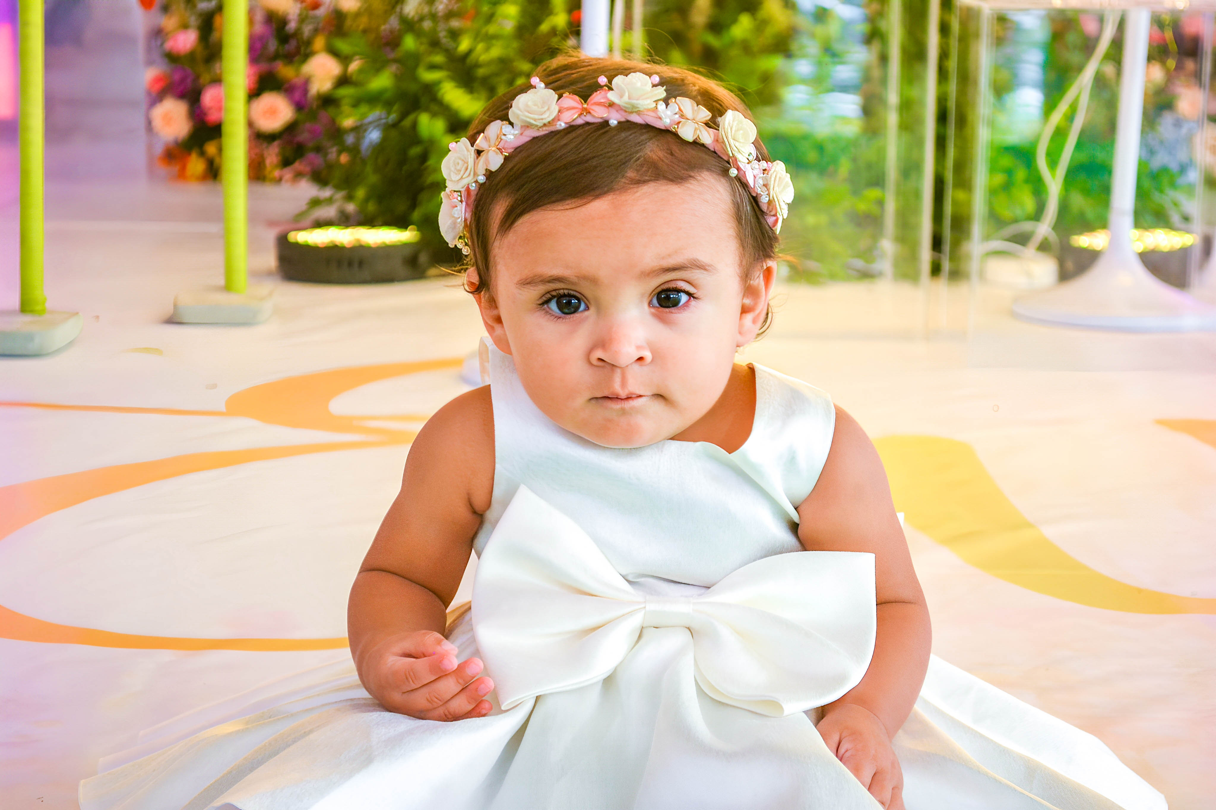 Bebê sentada em tapete verde, vestindo traje branco e posando em um ambiente com decoração floral e temática de fadas.