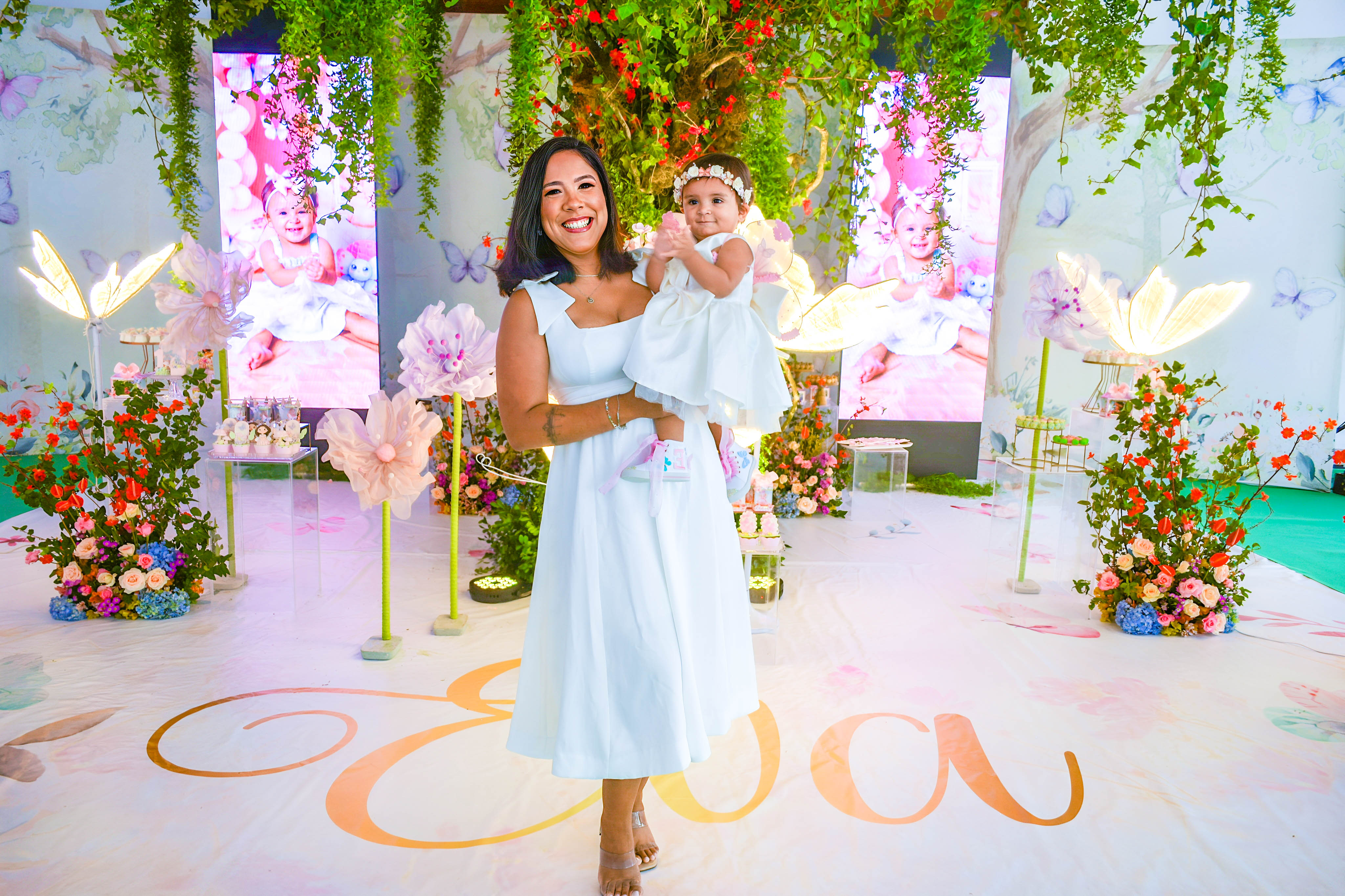 Duas mulheres fantasiadas de fadas em um ambiente decorado e florido, sorrindo enquanto a criança observa a cena.