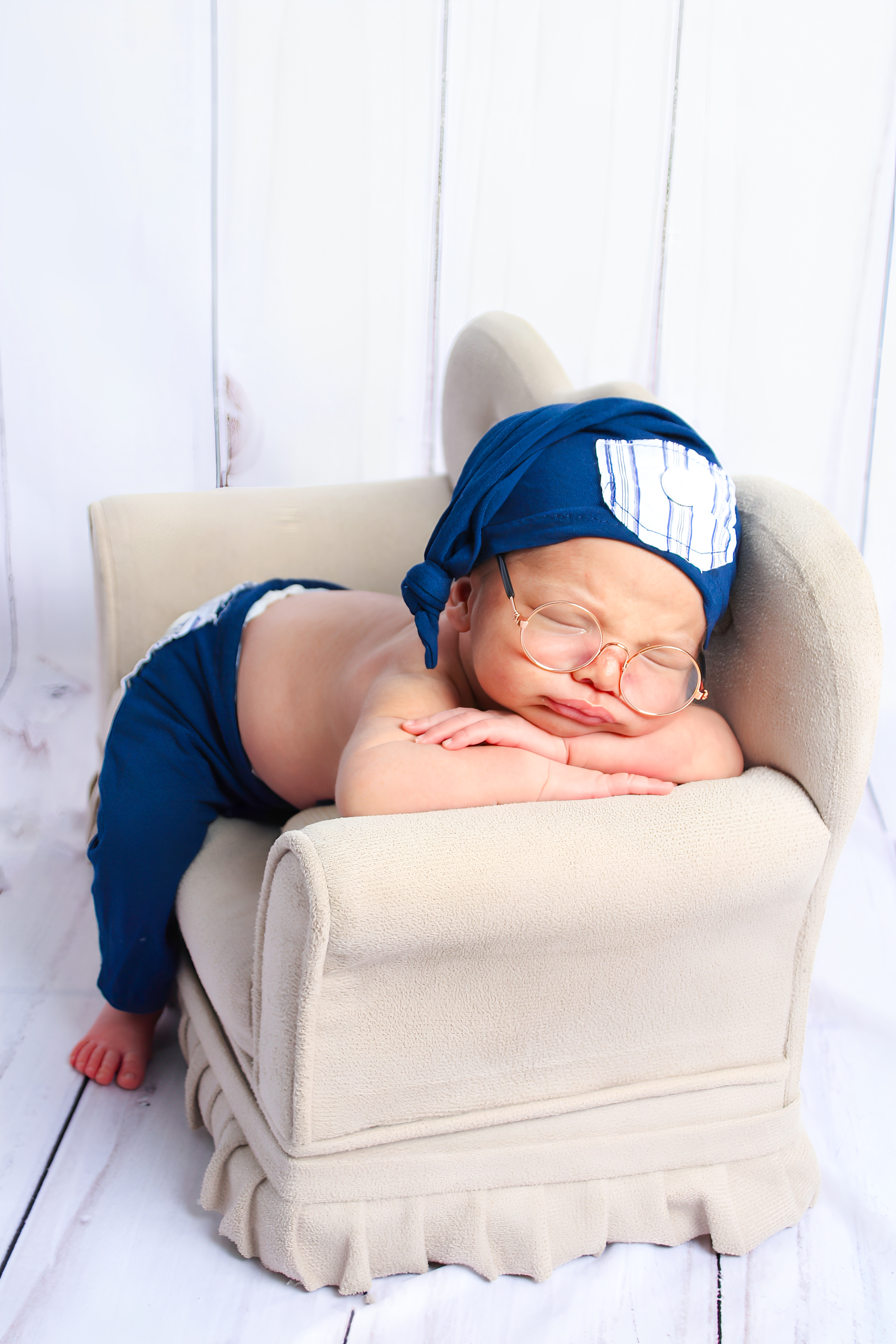 Recém-nascido dormindo em uma mini poltrona, usando touca e roupinha azul no ensaio newborn.