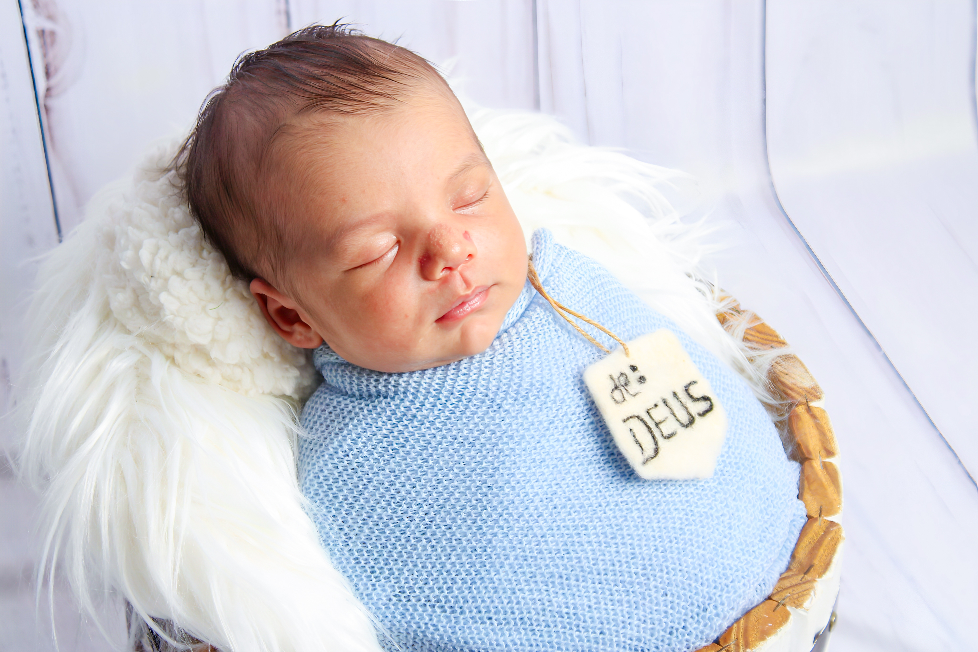 Bebê dormindo em roupinha azul com uma plaquinha 'É de Deus' no ensaio newborn.