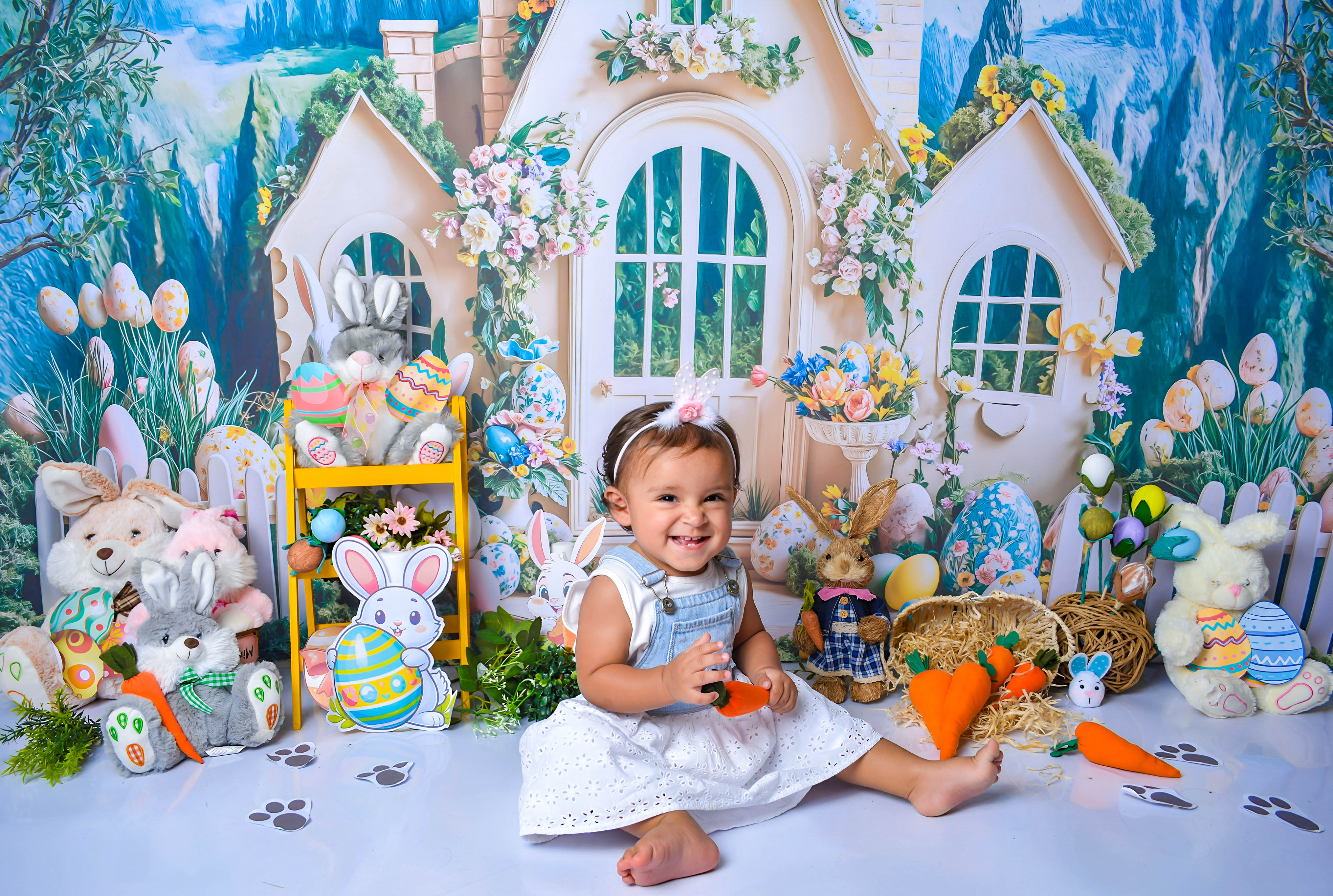 Bebê com vestido branco, sorrindo em um cenário de Páscoa com coelhos e cenouras.