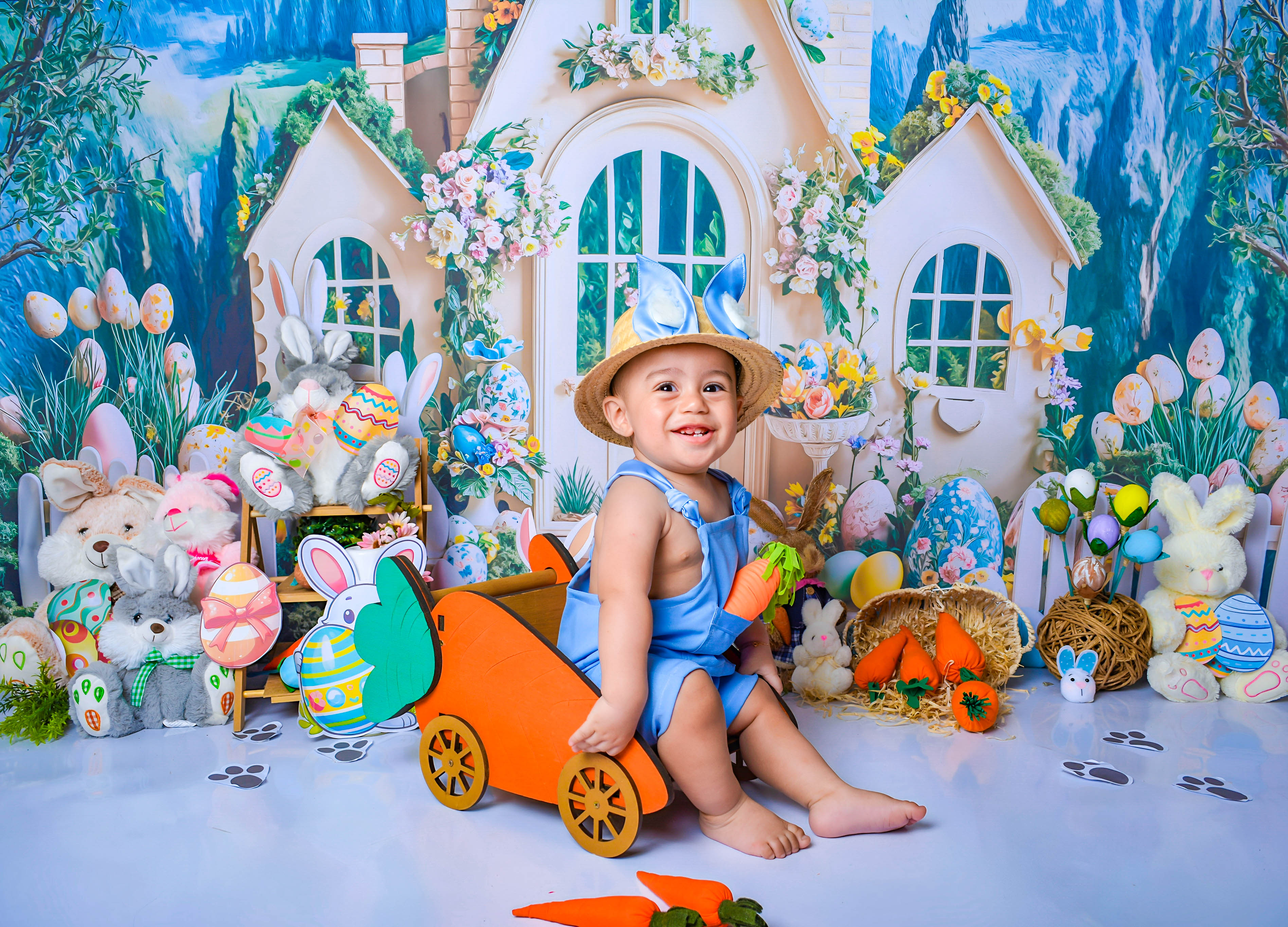 Menino usando chapéu e macacão azul, posando ao lado de um carrinho decorativo de cenoura, em um ensaio de Páscoa.